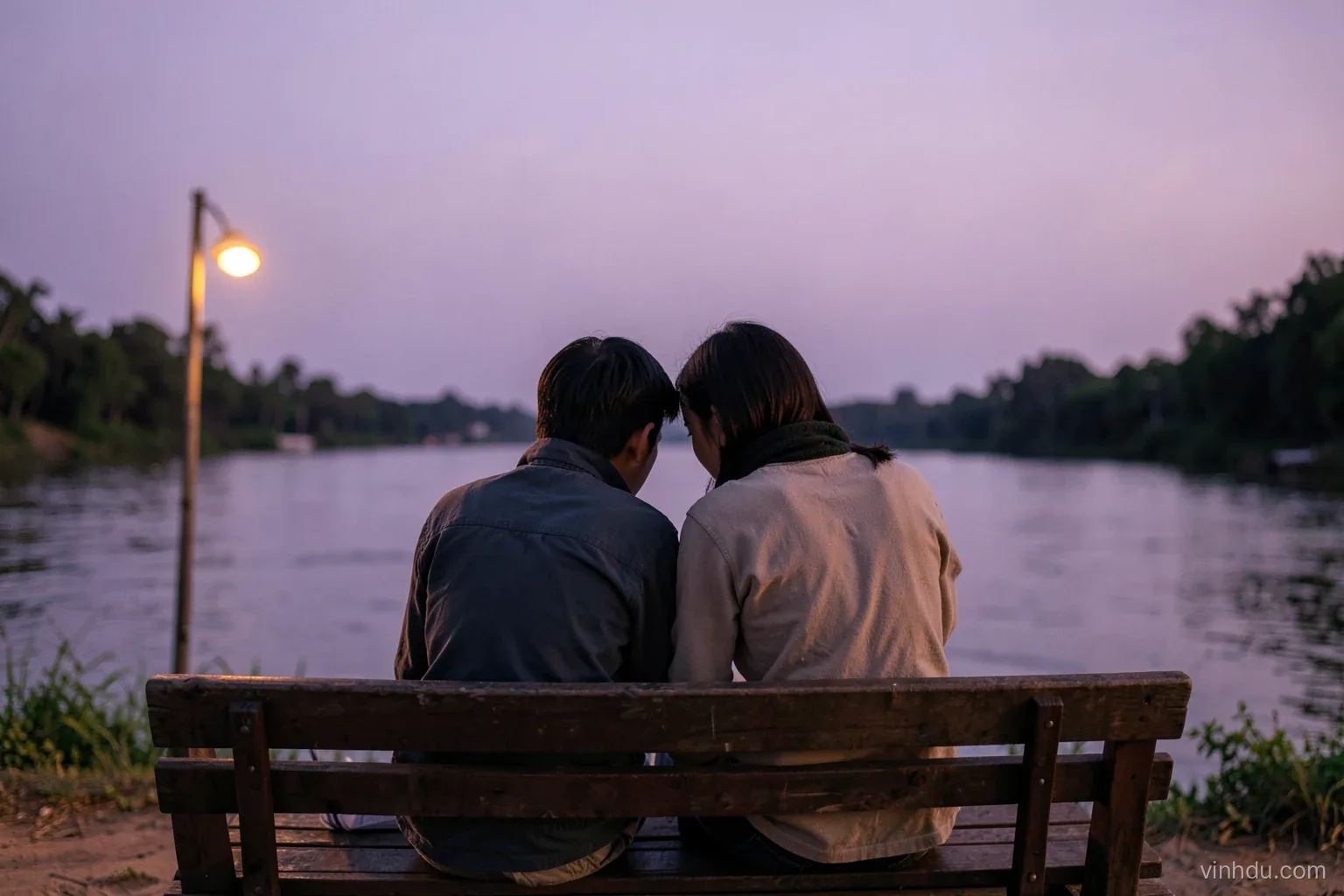 Two Vietnamese friends seen from behind sitting close on a quiet riverside bench at early evening, one leaning in listening, soft lilac sky and warm lamp light