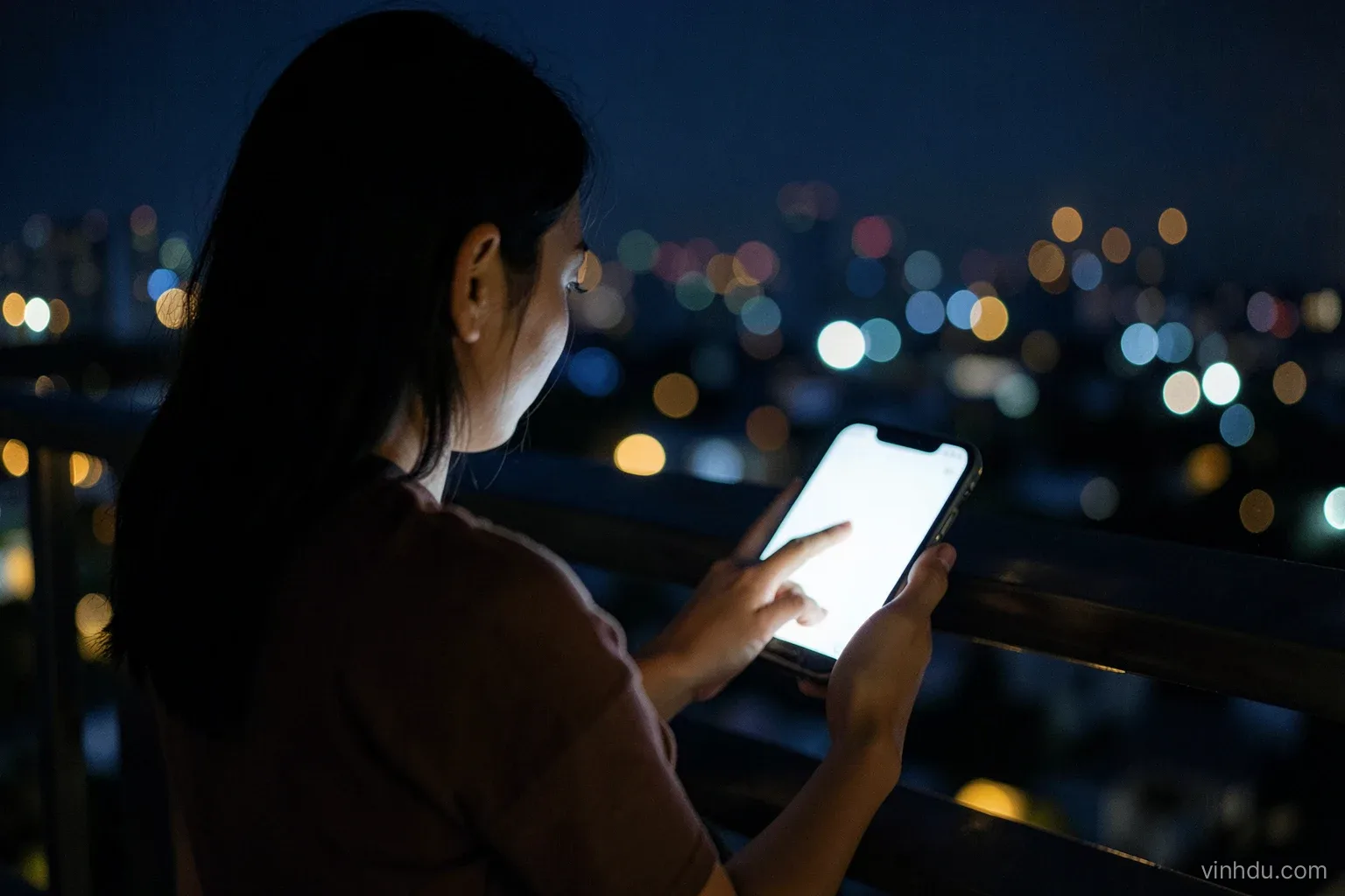 A young Vietnamese woman seen from behind sitting on a balcony at night, pausing with her thumb over a blank glowing phone, city lights softly blurred, deep blue and gold